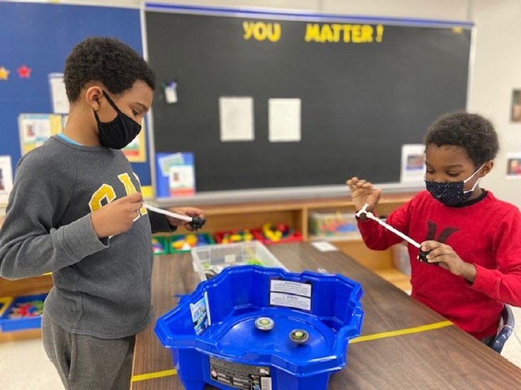 An image of two boys in masks playing with bey blades at a table